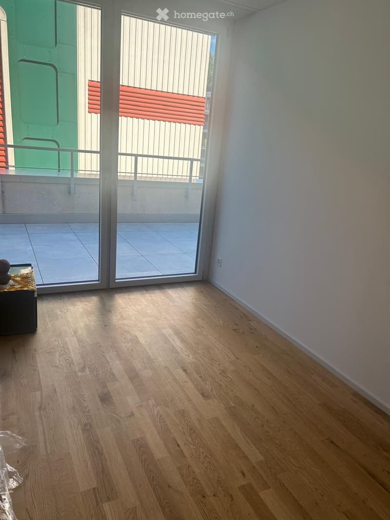 A small, unfurnished room with light wooden flooring and white walls, featuring a sliding glass door leading to a tiled balcony area.