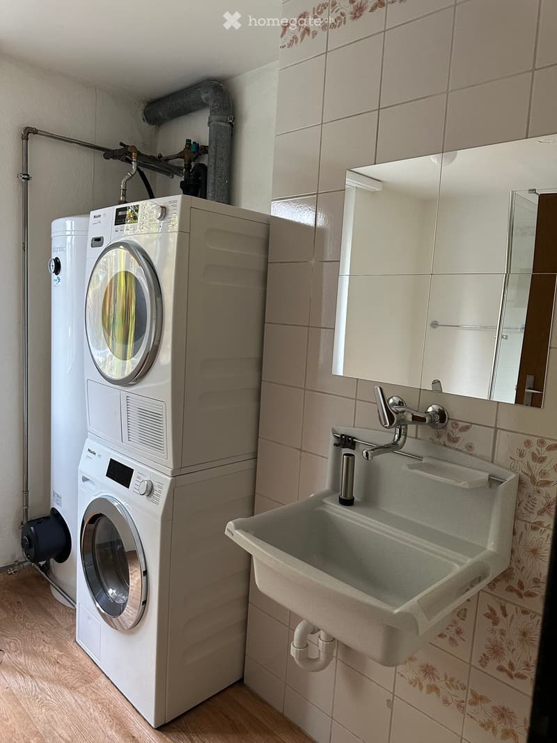 Bathroom with a washing machine, sink, and mirror. The walls are tiled with a floral pattern, and the floor is wooden.