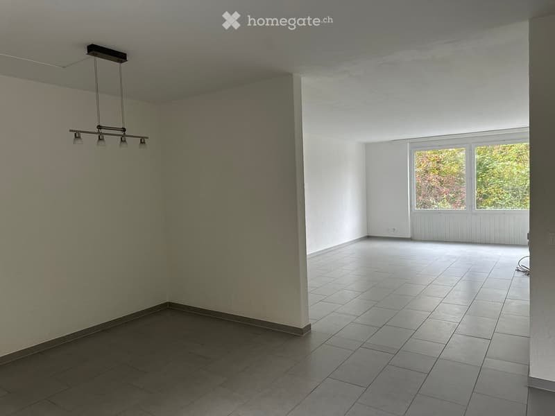 Empty room with tiled flooring, white walls, and large windows offering a view of greenery outside. A modern light fixture hangs from the ceiling.