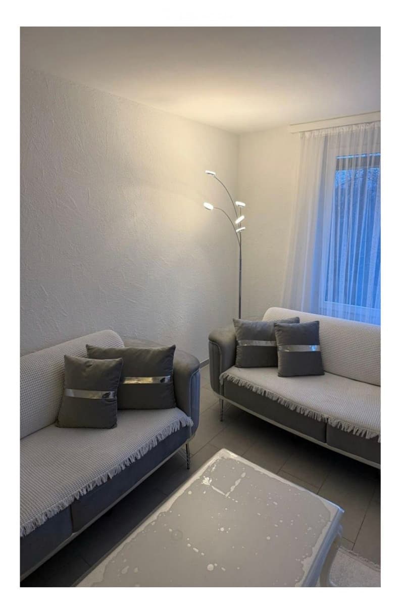 Living room with two sofas, a coffee table, and a floor lamp. The room features a large window with sheer curtains and a neutral color palette.
