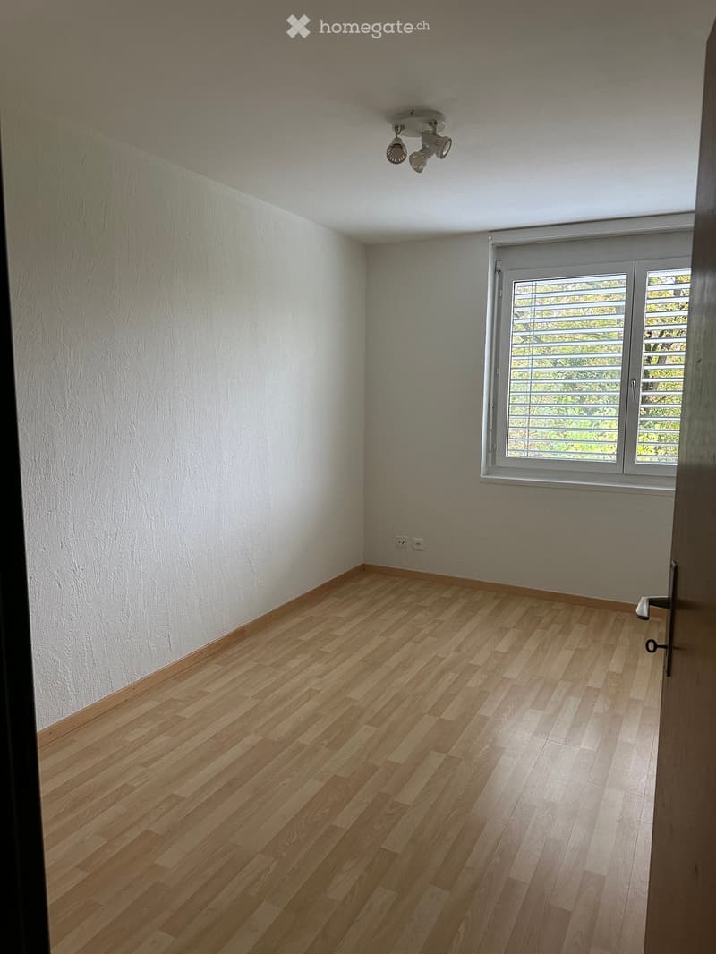 Empty room with wooden flooring, white walls, and a large window with blinds. A ceiling light fixture is visible.