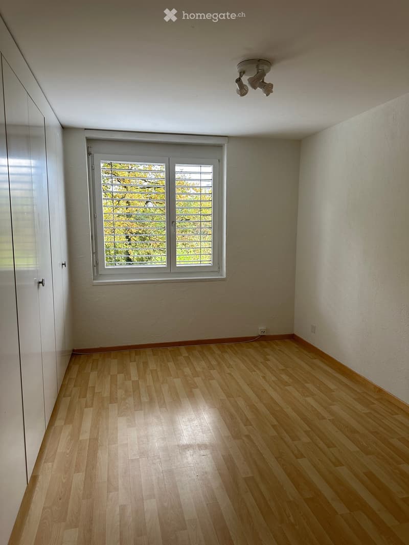 Empty room with light wooden flooring, white walls, and a large window with blinds. The ceiling features a modern light fixture.