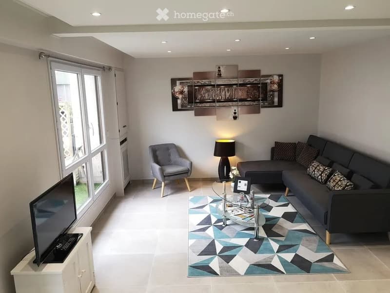 Modern living room with a grey sectional sofa, armchair, glass coffee table, TV stand, floor lamp, and a geometric patterned rug.