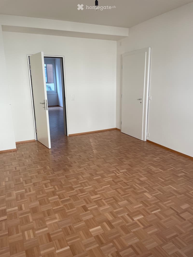 Empty room with wooden herringbone flooring, white walls, and two doors, one leading to another room.