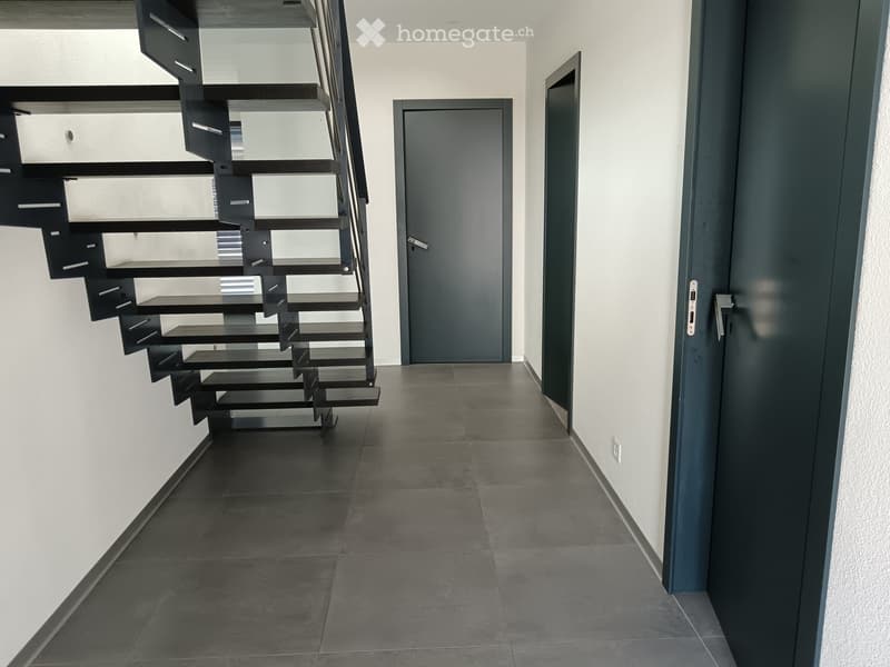 Modern hallway with dark tiled floor, white walls, and a sleek black staircase leading upwards. Two doors are visible, one on each side of the hallway.