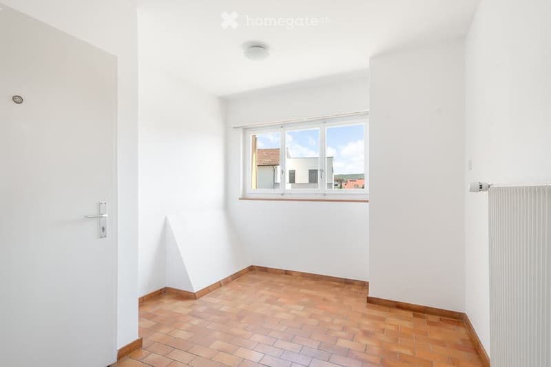 Empty room with white walls, wooden tiled floor, and a window with a view of a building outside.