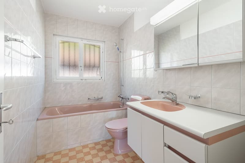 Bathroom with a pink bathtub, toilet, sink, and mirror. The walls are tiled in light beige, and the floor has a checkered pattern. A window with frosted glass is present.