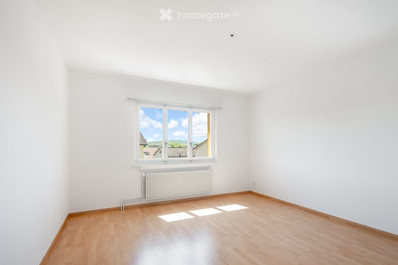 Empty room with white walls, wooden flooring, large window with view, radiator below window.