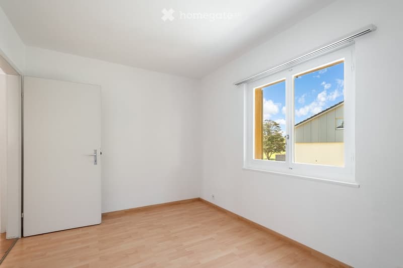Empty room with white walls, wooden flooring, and large windows showing a view of the sky and a tree outside.