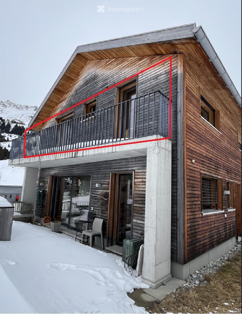 Two-story wooden house with a balcony, large windows, and a modern design, set in a snowy landscape with mountains in the background.
