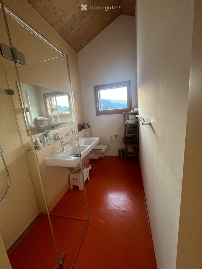 Bathroom with red flooring, white sink, toilet, mirror, and small shelf. Features a window with a scenic view.