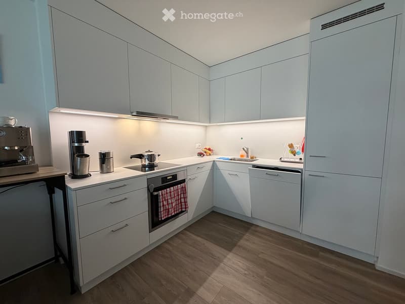 Modern kitchen with white cabinets, wooden flooring, and stainless steel appliances including a coffee maker, oven, and dishwasher. Countertop features a sink, faucet, and small decorative items.