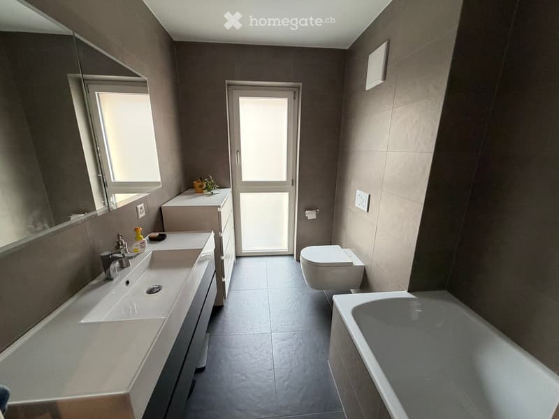 Modern bathroom with double sink, toilet, bathtub, and large mirror. Features grey tiles and a window providing natural light.