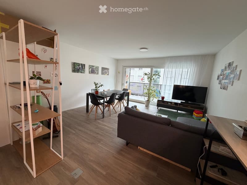 Open-plan living and dining area with a grey sofa, dining table with chairs, a guitar, a shelf with books and decorative items, and a television on a stand.