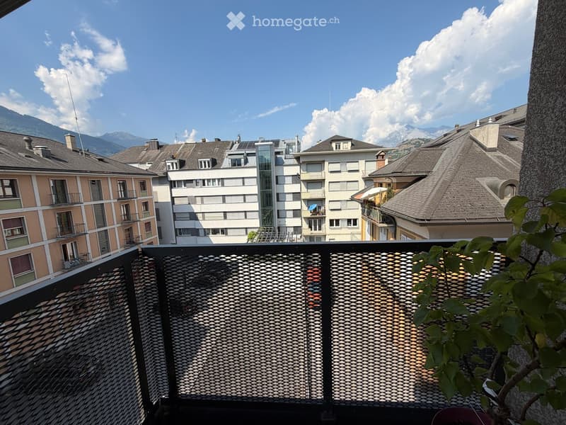 Balcony with metal railing, overlooking a cityscape with buildings and mountains in the background.