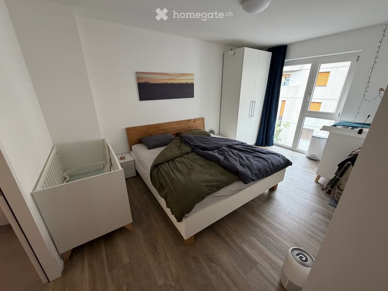Cozy bedroom with a wooden bed, nightstand, radiator, wardrobe, and a fan on the floor. The room features wooden flooring and large windows with dark curtains.