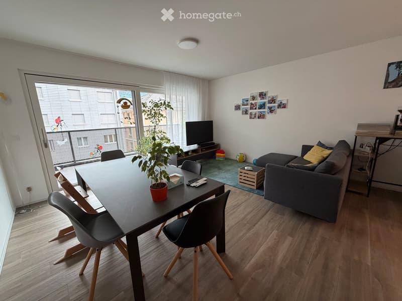 Modern living room with a grey sofa, a black dining table with four chairs, a potted plant, a television, and a small balcony with a view.