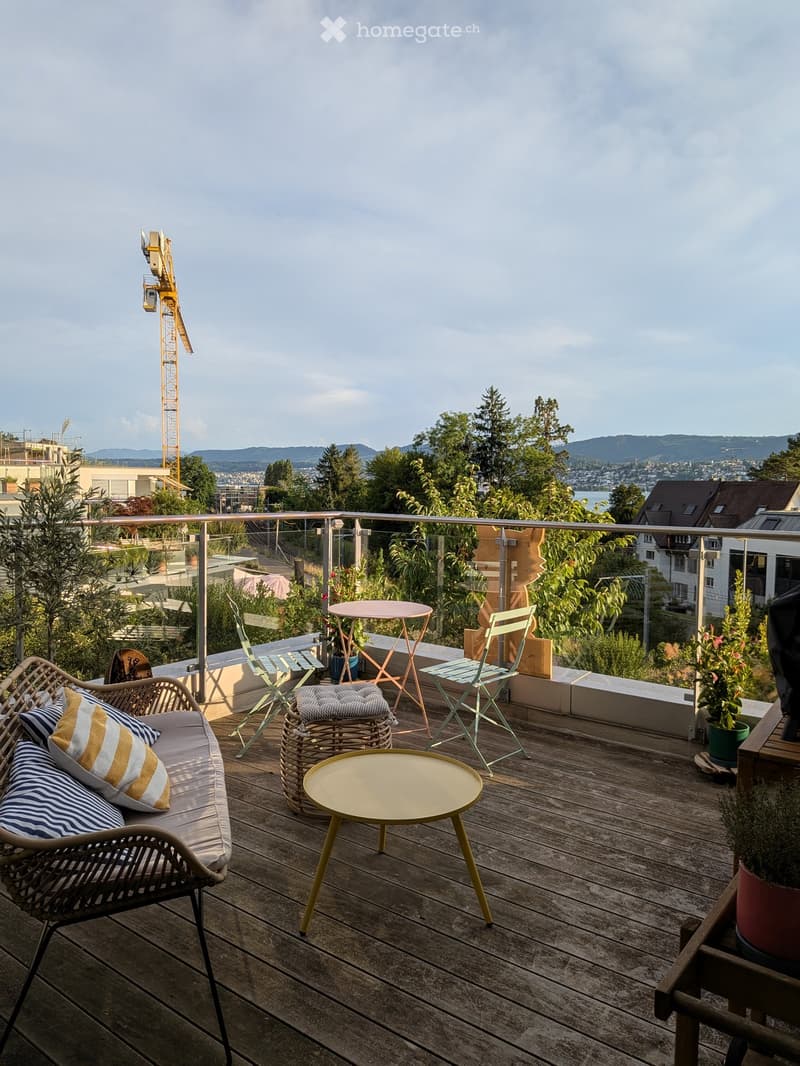 Wooden deck with a sofa, small tables, chairs, and potted plants, overlooking a cityscape with trees and a crane in the distance.