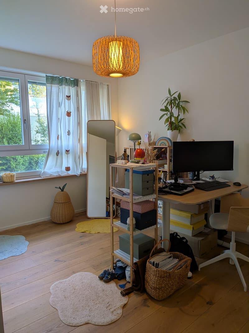 A well-lit room with wooden flooring, a large window with curtains, a desk with a computer setup, a shelf with storage boxes, a mirror, and decorative items including a plant and a wicker basket.