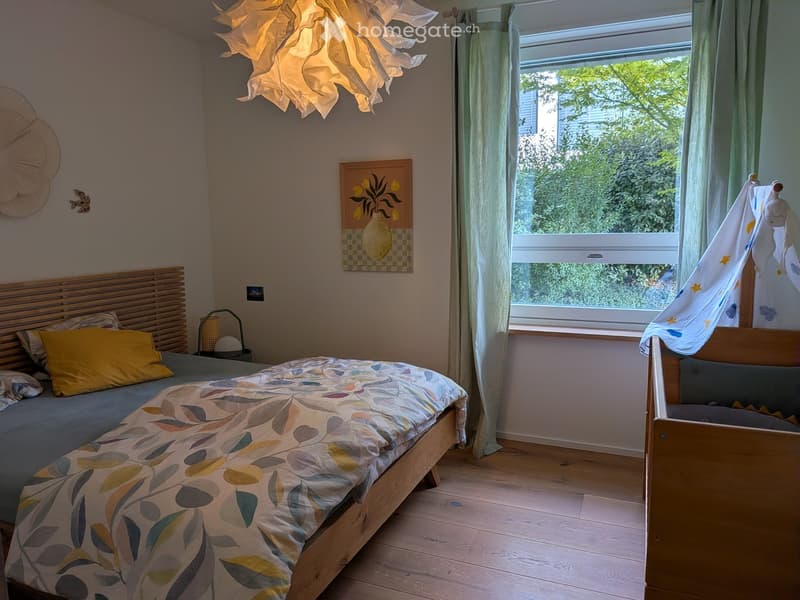 Cozy bedroom with a wooden bed, colorful bedding, a nightstand with a lamp, a large window with green curtains, a decorative picture frame, and a small baby crib with a toy tent.