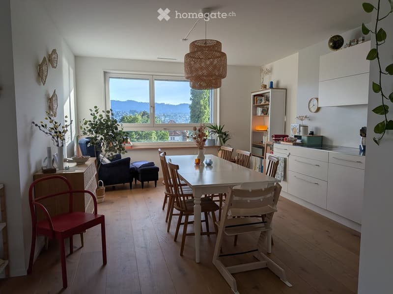 Bright, open-plan living and dining area with wooden flooring, large windows offering a scenic view, a dining table with chairs, a sofa, a stool, a kitchen area with cabinets, a bookshelf, and decorative plants.