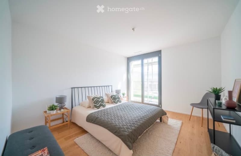 Bright, modern bedroom with a large bed, nightstand, lamp, chair, small table with plant, and picture frame on the wall. The room features wooden flooring and large windows with a view of the outdoor area.