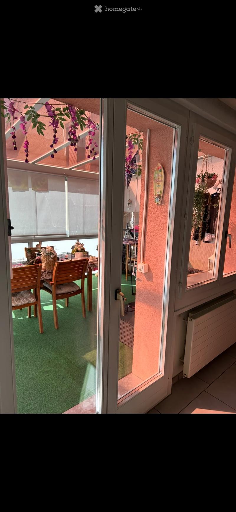 Balcony with green artificial turf, wooden chairs, small table, and flower pots. Visible through glass doors from the interior.