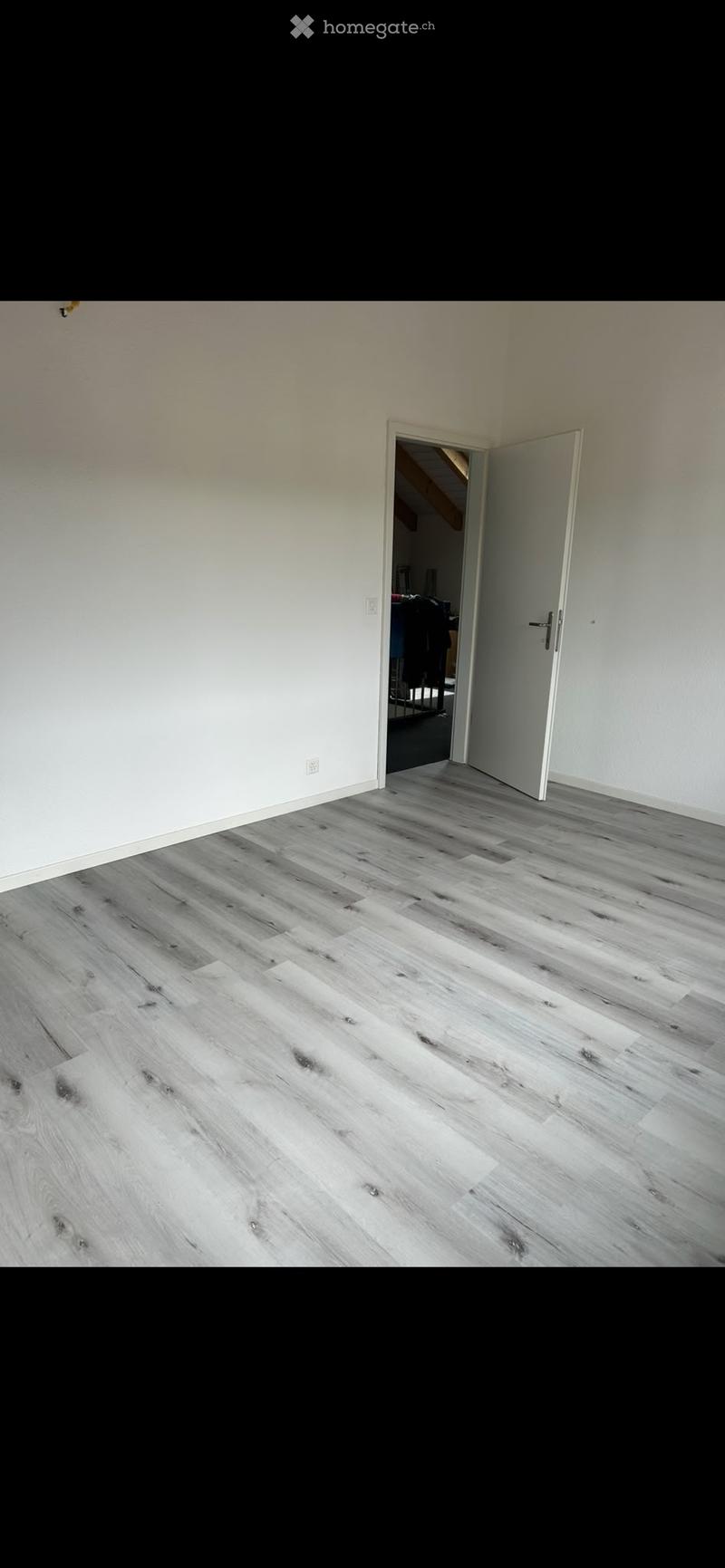 Empty room with light-colored walls and a wooden floor, featuring an open doorway leading to another room.