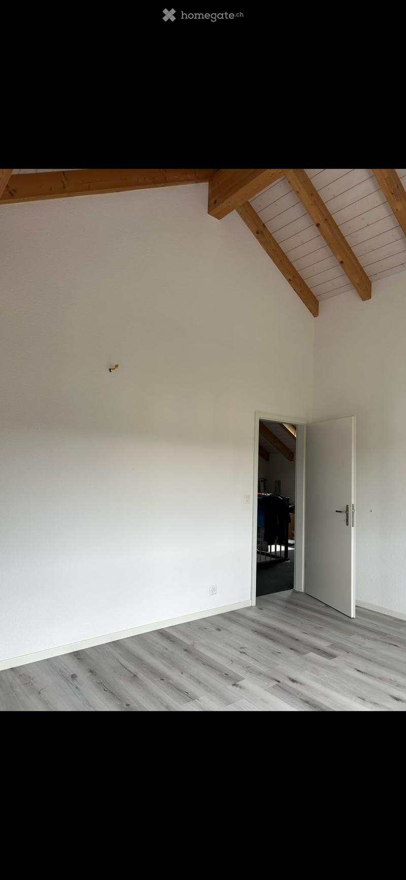 Empty room with light grey wooden flooring and white walls, featuring exposed wooden beams on the ceiling and an open doorway leading to another room.