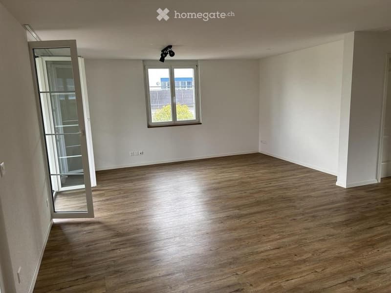 Empty room with wooden flooring, white walls, and a large window with a view of the outside. The room features a sliding glass door leading to a balcony area.