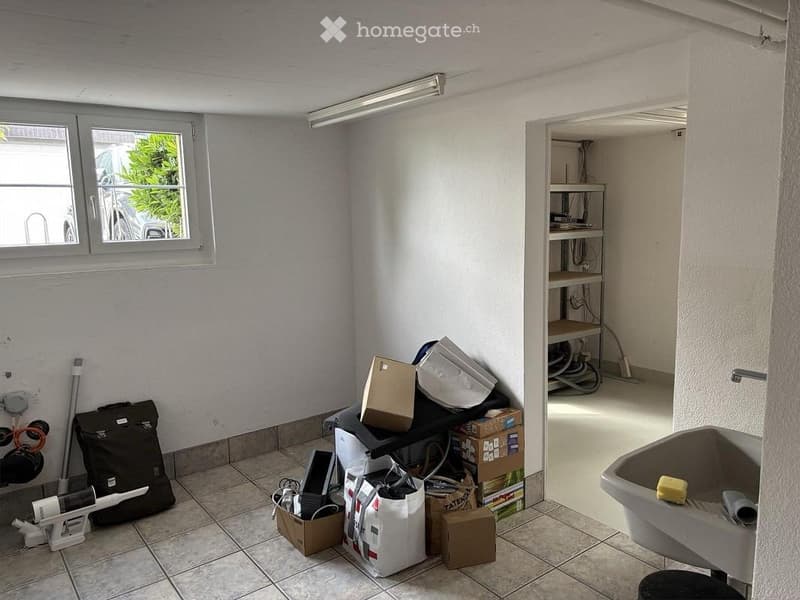 Empty room with tiled floor, white walls, and a window. Various items such as boxes, bags, and a shelf are present, but the room itself is unfurnished.