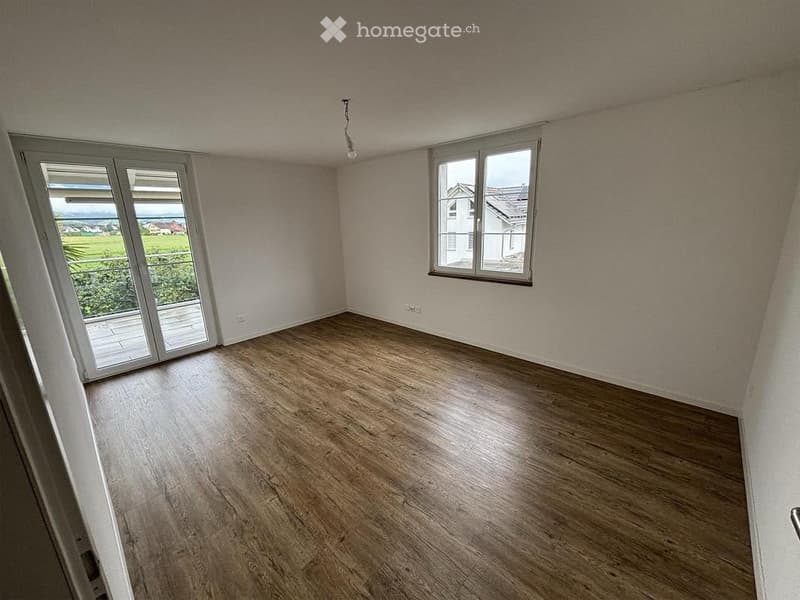 Empty room with wooden flooring, white walls, large window, and sliding glass door leading to a balcony.