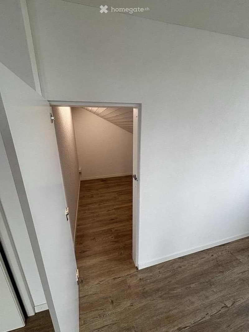 Empty room with white walls and wooden flooring, visible through an open doorway.