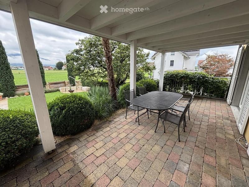 Brick-paved balcony with a table and chairs, overlooking a landscaped garden with trees and a lawn.