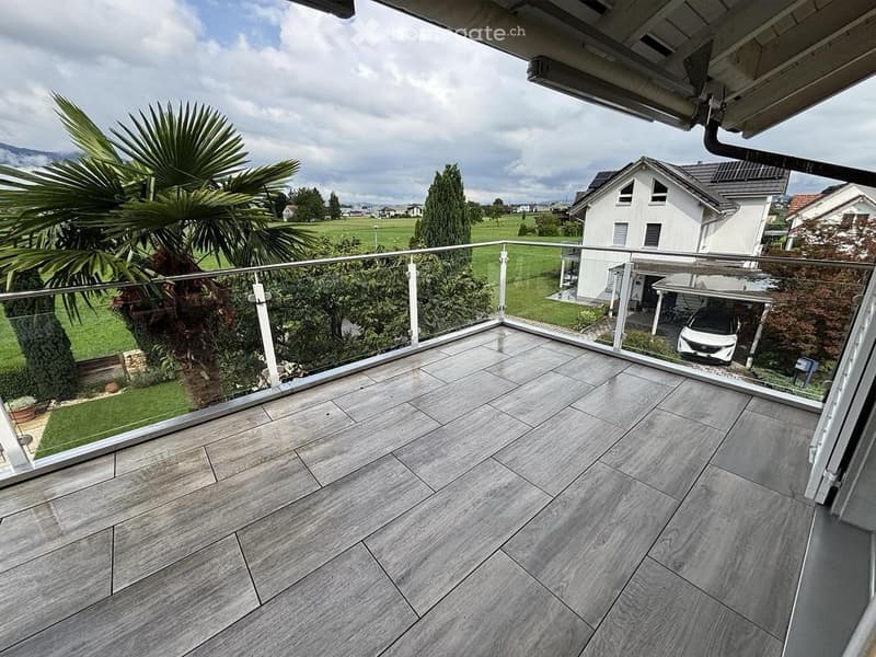 Balcony with modern glass railing, tiled floor, and view of a green landscape with houses and trees.