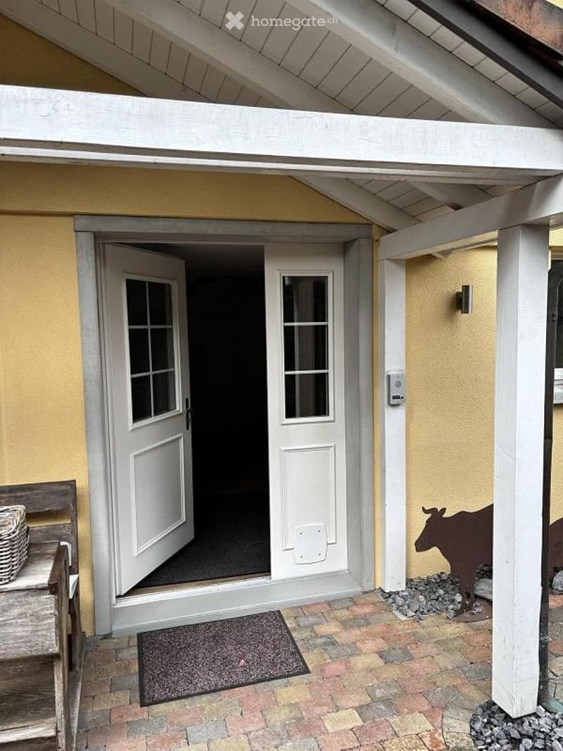 Entrance area with double doors leading inside, a doormat, and a small wooden bench with a woven basket on it. The walls are painted yellow, and there is a decorative cow statue on the right side.