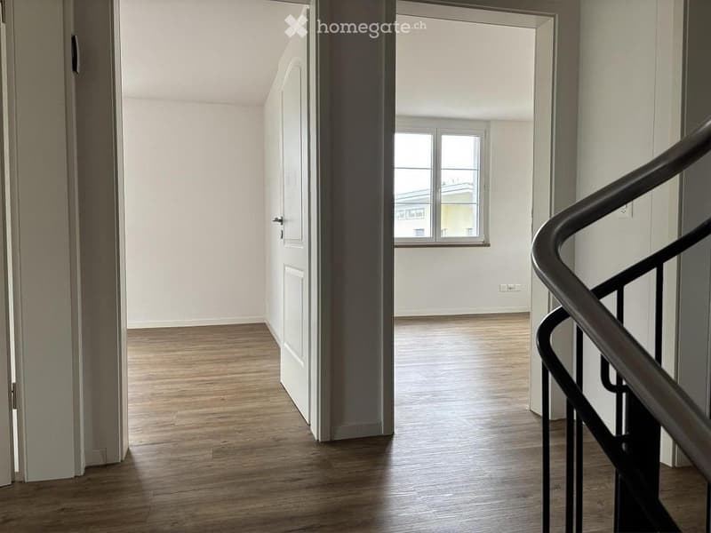 Empty hallway with wooden flooring, white walls, and a staircase leading upwards. Natural light streams in from a window at the end of the corridor.