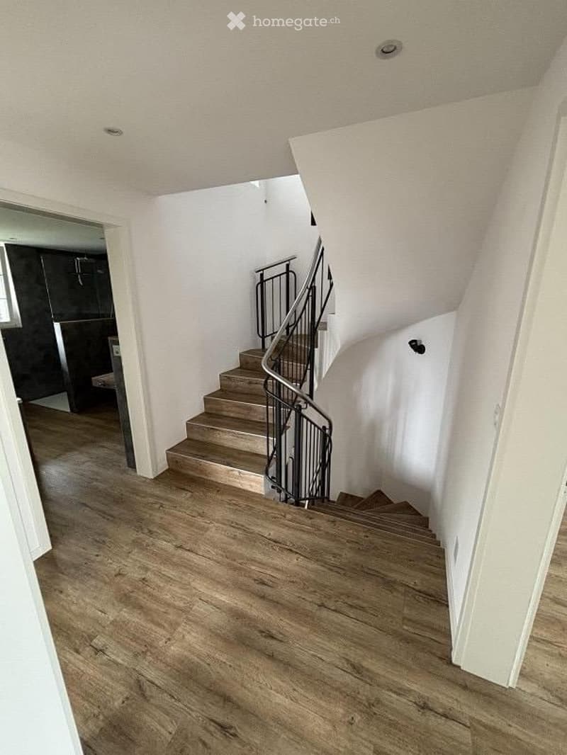 Spacious hallway with wooden flooring and a staircase leading upwards, featuring a modern metal railing and white walls.