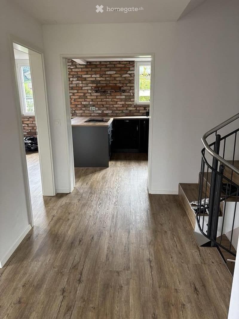 Open-plan area with wooden flooring, modern kitchen area with dark cabinets and a central island, exposed brick wall, and a staircase leading to an upper level.