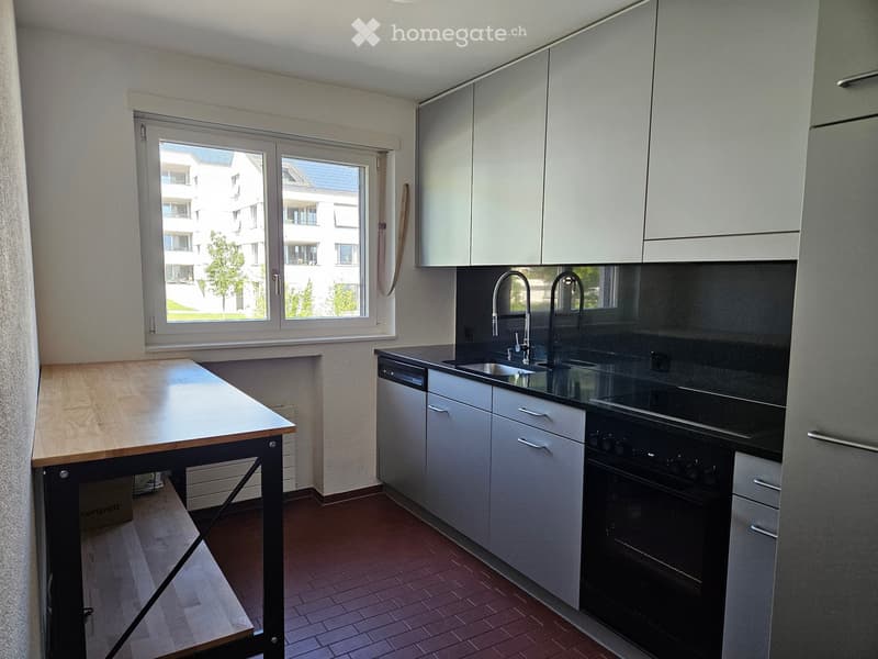 Modern kitchen with white cabinets, a wooden table, a sink with a faucet, an oven, and a dishwasher. The room has a large window offering a view of the building exterior.
