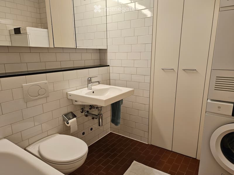 Modern bathroom with white tiles, a toilet, a sink with a chrome faucet, a mirror, and a towel hanging on the side.