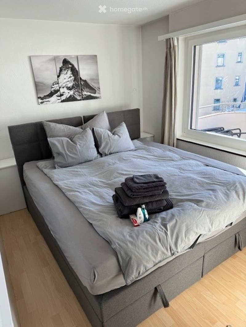 double bed with a gray mattress, headboard, and pillows, wooden floors, large windows with curtain, nightstand, picture frame, towels, and a bottle on the bed