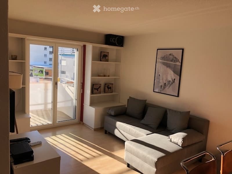 Sliding glass door leading to a balcony, white wall, gray couch, gray ottoman, white wooden table, shelf, painting, remote