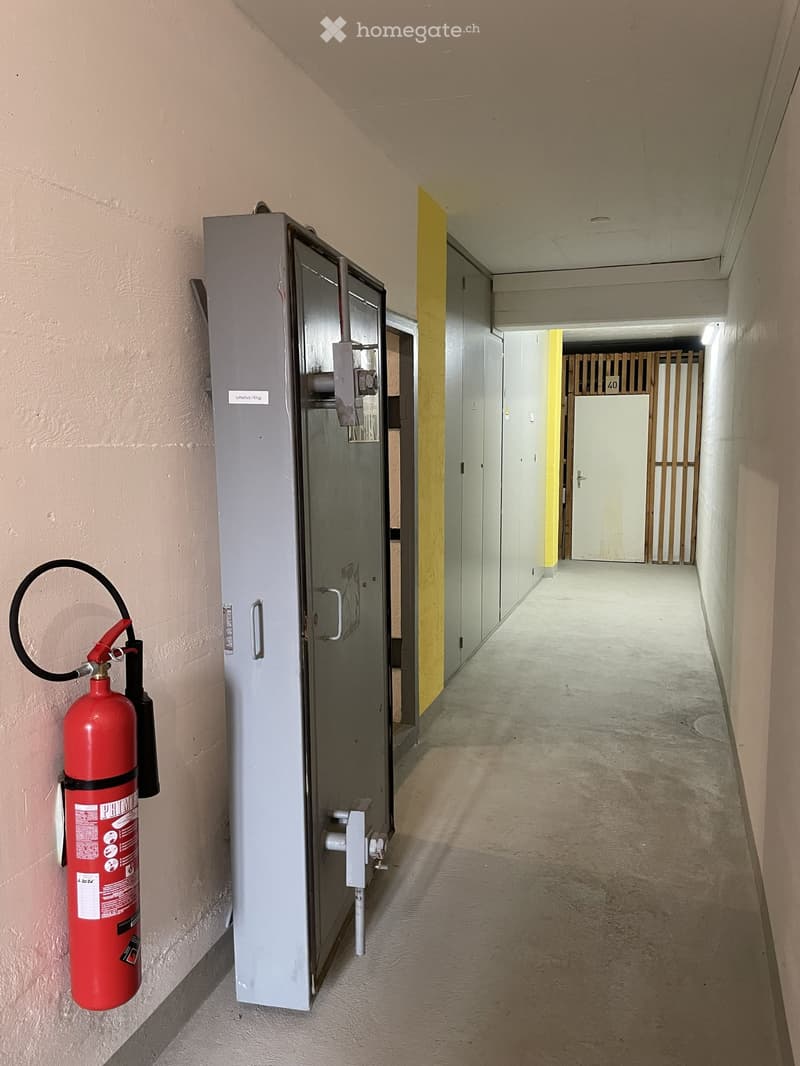 Empty hallway with a fire extinguisher mounted on the wall and a grey electrical panel next to it. The hallway has beige walls and a concrete floor, with a yellow accent wall visible in the distance.