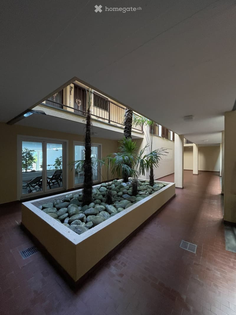 Indoor courtyard with a raised planter bed containing rocks and small trees, surrounded by a tiled floor and walls, with a balcony visible above.