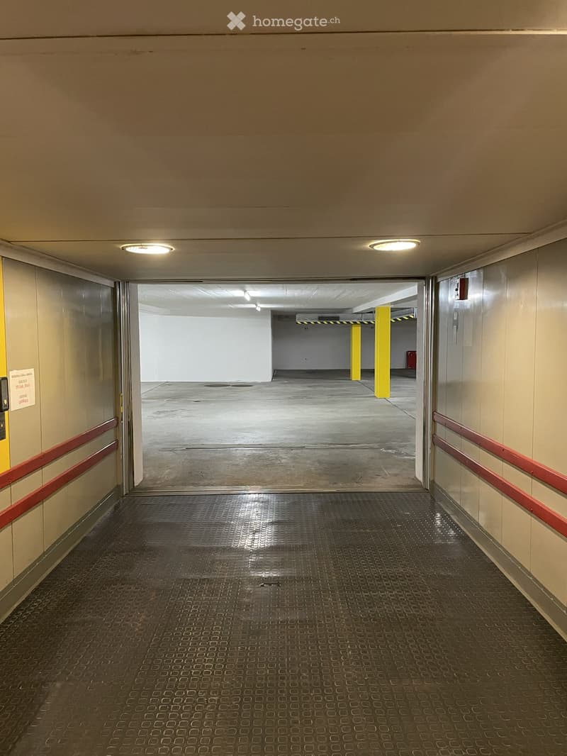 A clean, well-lit hallway with beige walls, a metal floor, and red handrails. An open doorway leads to a parking area with yellow pillars and a concrete floor.