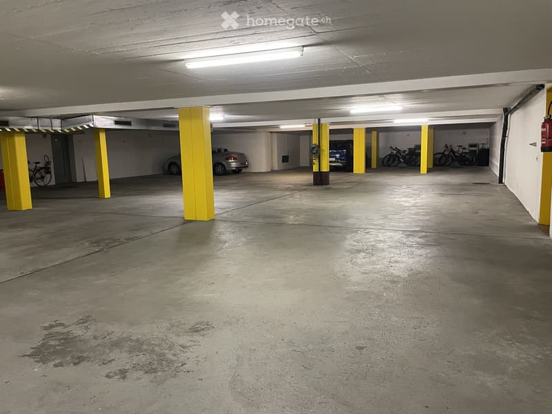 Empty underground parking area with concrete floors, yellow support pillars, and a few parked vehicles and bicycles in the background.