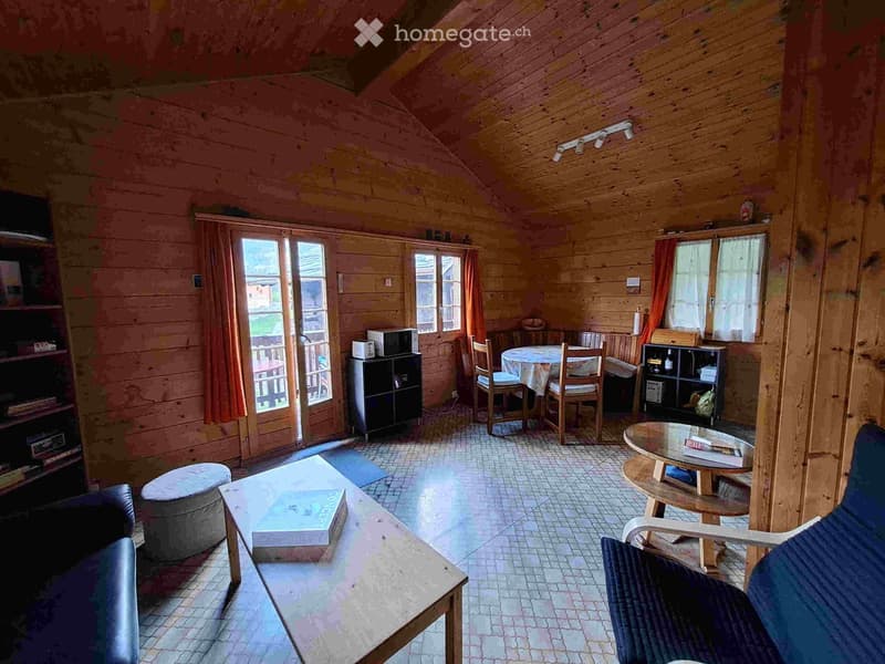 A cozy, wooden-paneled living room with a slanted ceiling, featuring a sofa, coffee table, dining area with chairs and table, and shelves with various items.