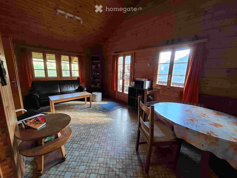 A cozy living room with wooden beams, large windows, a black leather sofa, a wooden coffee table, a round wooden table with chairs, and a bookshelf.
