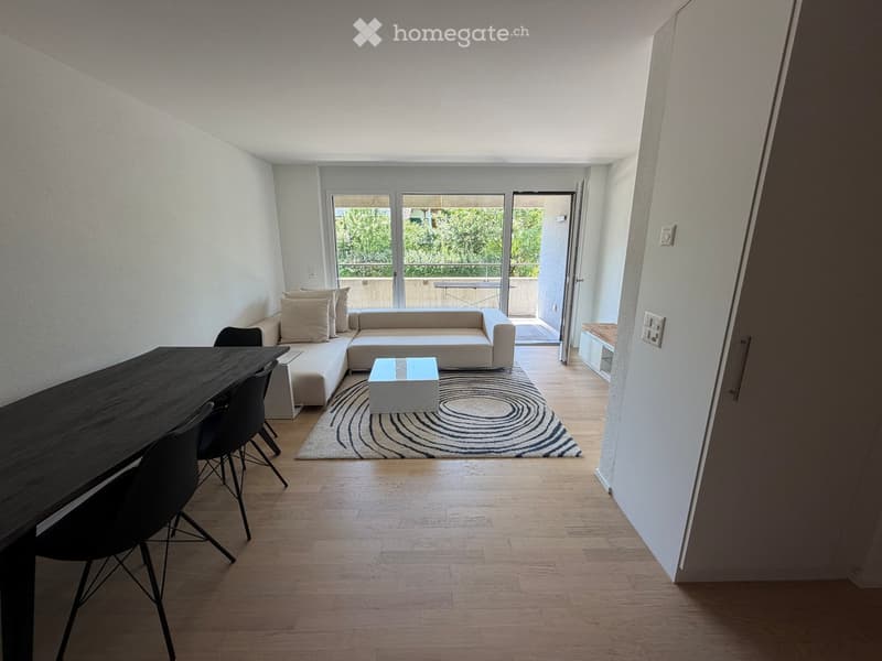 Modern living and dining area with a white sectional sofa, coffee table, dining table, and chairs. The space features light wooden flooring, a patterned rug, and large windows leading to a balcony.